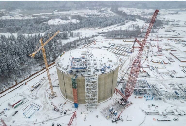 Vue aérienne d’un vaste chantier industriel enneigé, avec un grand réservoir cylindrique en béton en construction, entouré de grues, d’équipements et de zones de stockage, au milieu d’un paysage forestier hivernal.