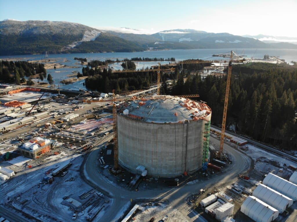 Grand réservoir cylindrique de stockage en construction sur un site industriel portuaire entouré de grues et de terrains enneigés, avec un plan d’eau et des montagnes en arrière-plan.