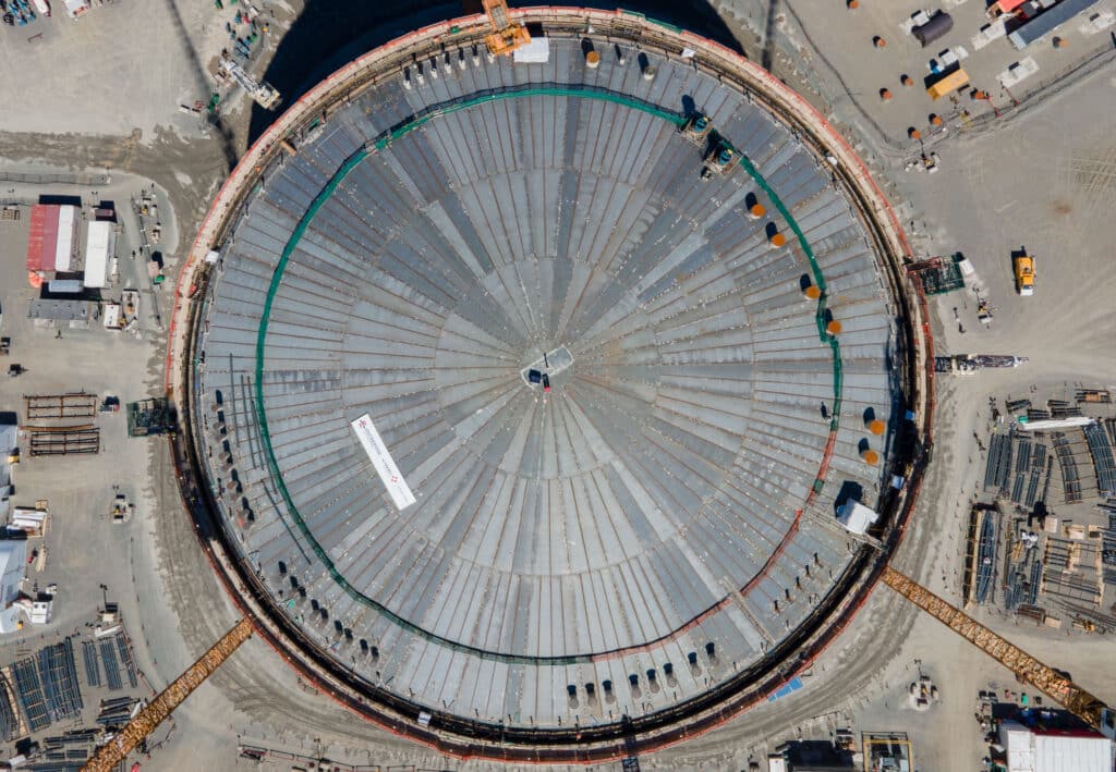 Vue aérienne d’un grand réservoir circulaire en construction sur un site industriel, avec grues, engins et matériaux disposés autour.